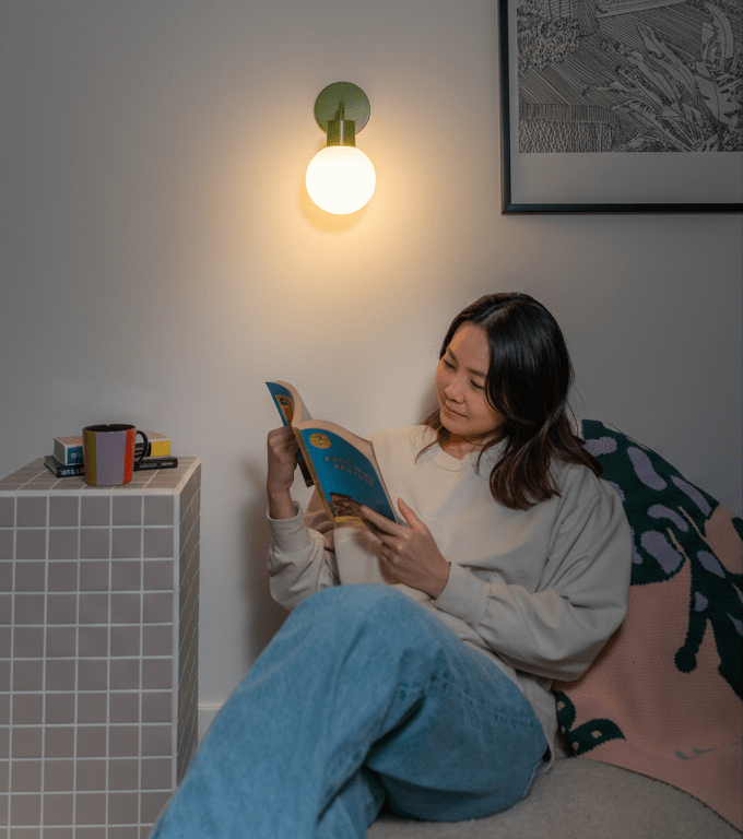 Woman reading a book in a cozy bedroom corner, illuminated by a Sage Green Poplight wall sconce.