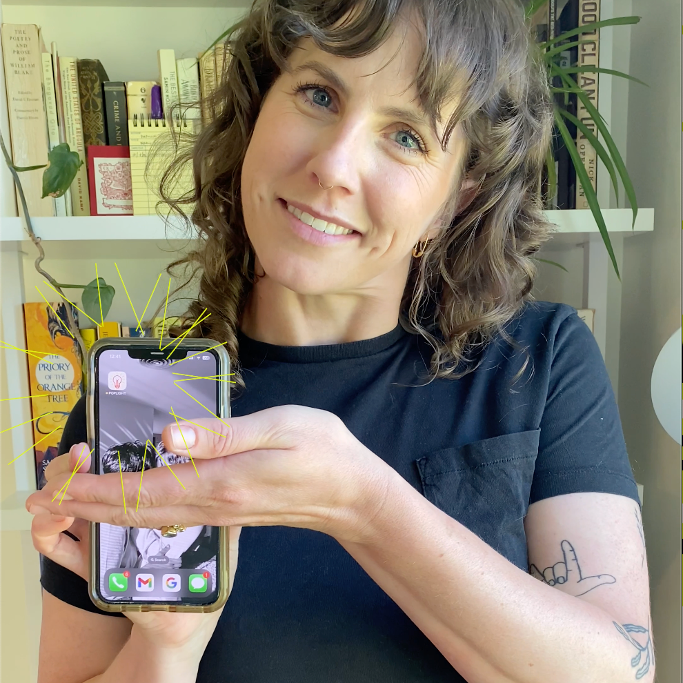 Smiling woman holding a smartphone displaying a creative app, surrounded by books and plants in bright space.