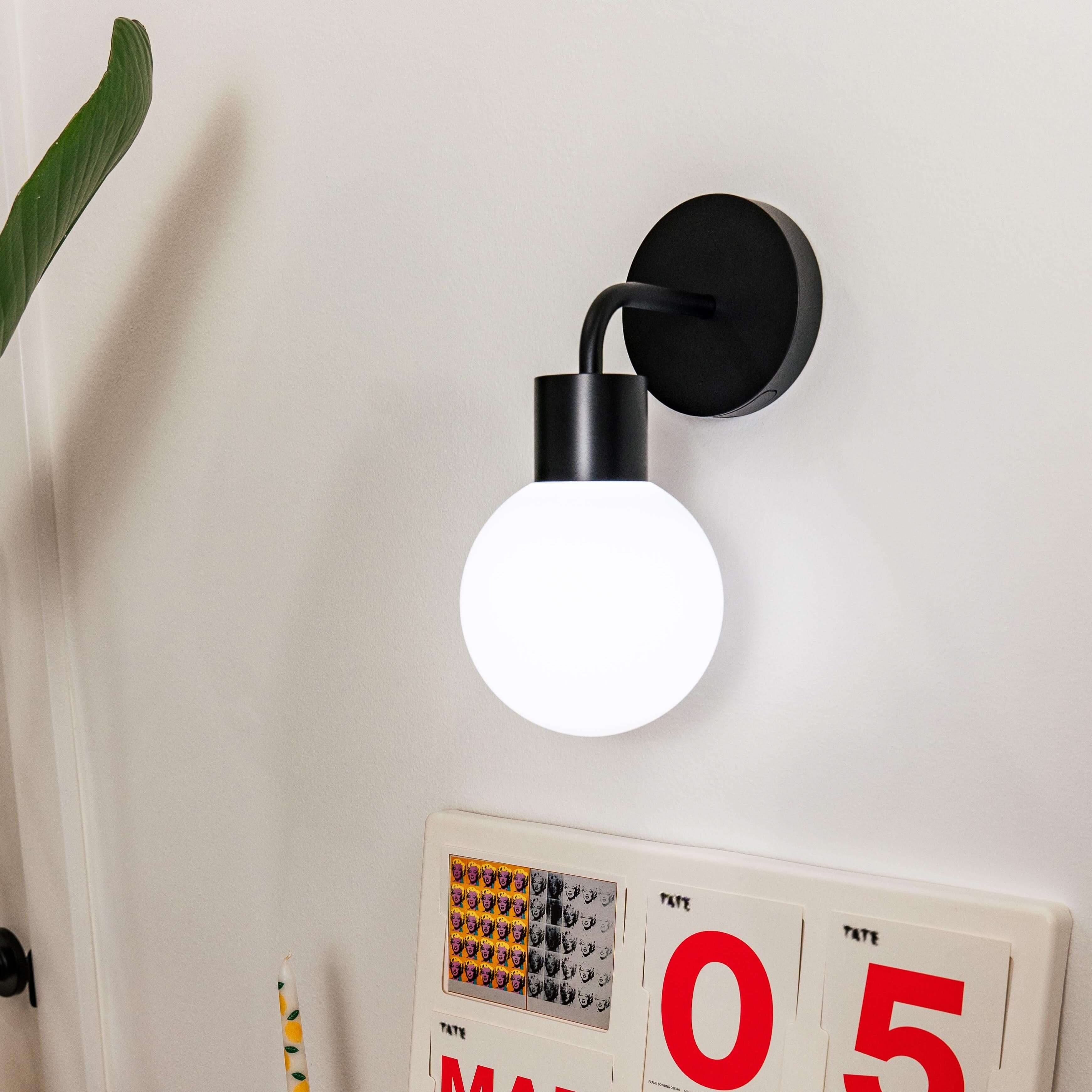 Modern rechargeable wall sconce with round bulb illuminating a stylish desktop calendar and decor setup on a white wall.