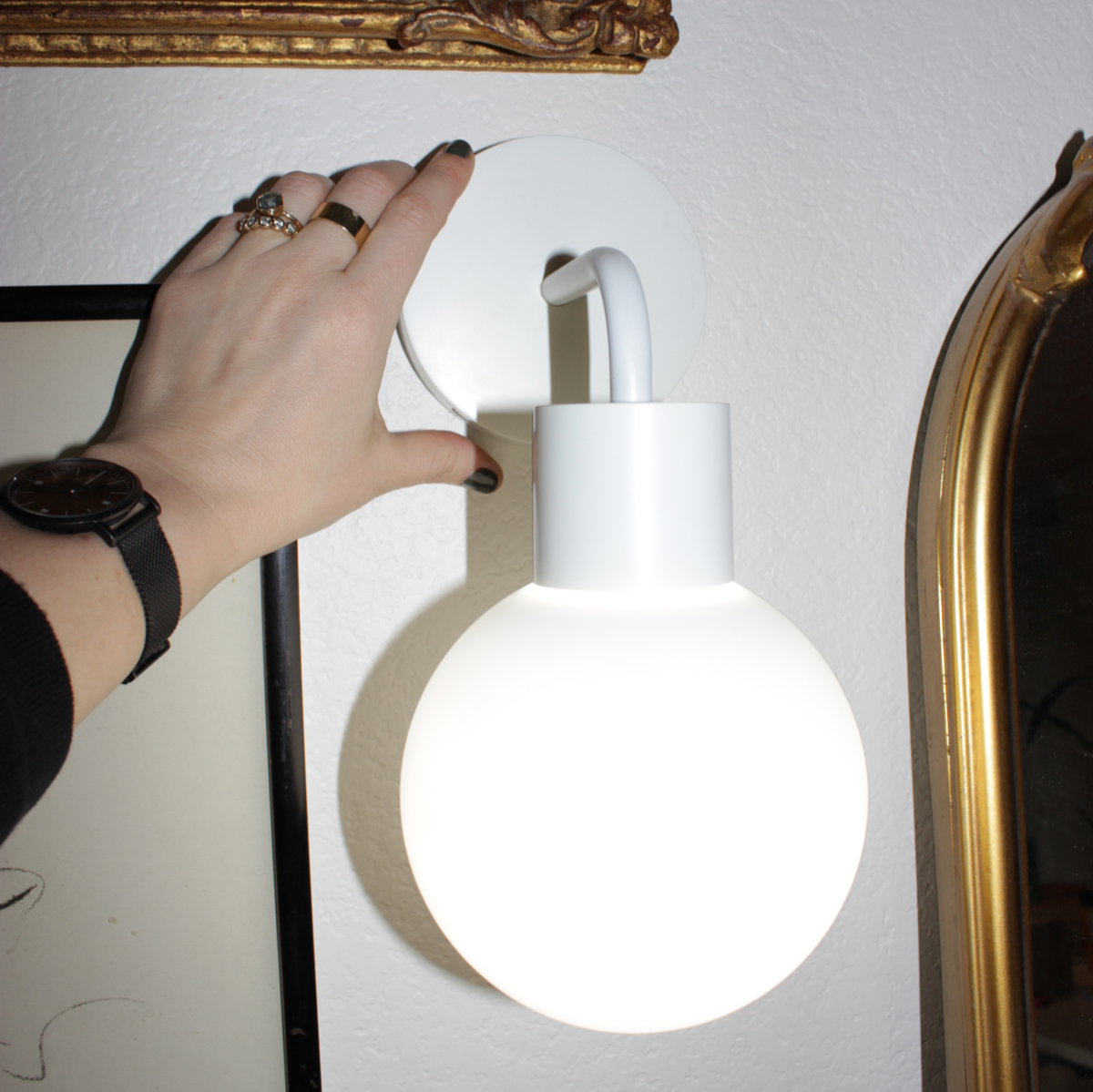 Hand adjusting modern white wall lamp with round bulb against textured wall and framed art.
