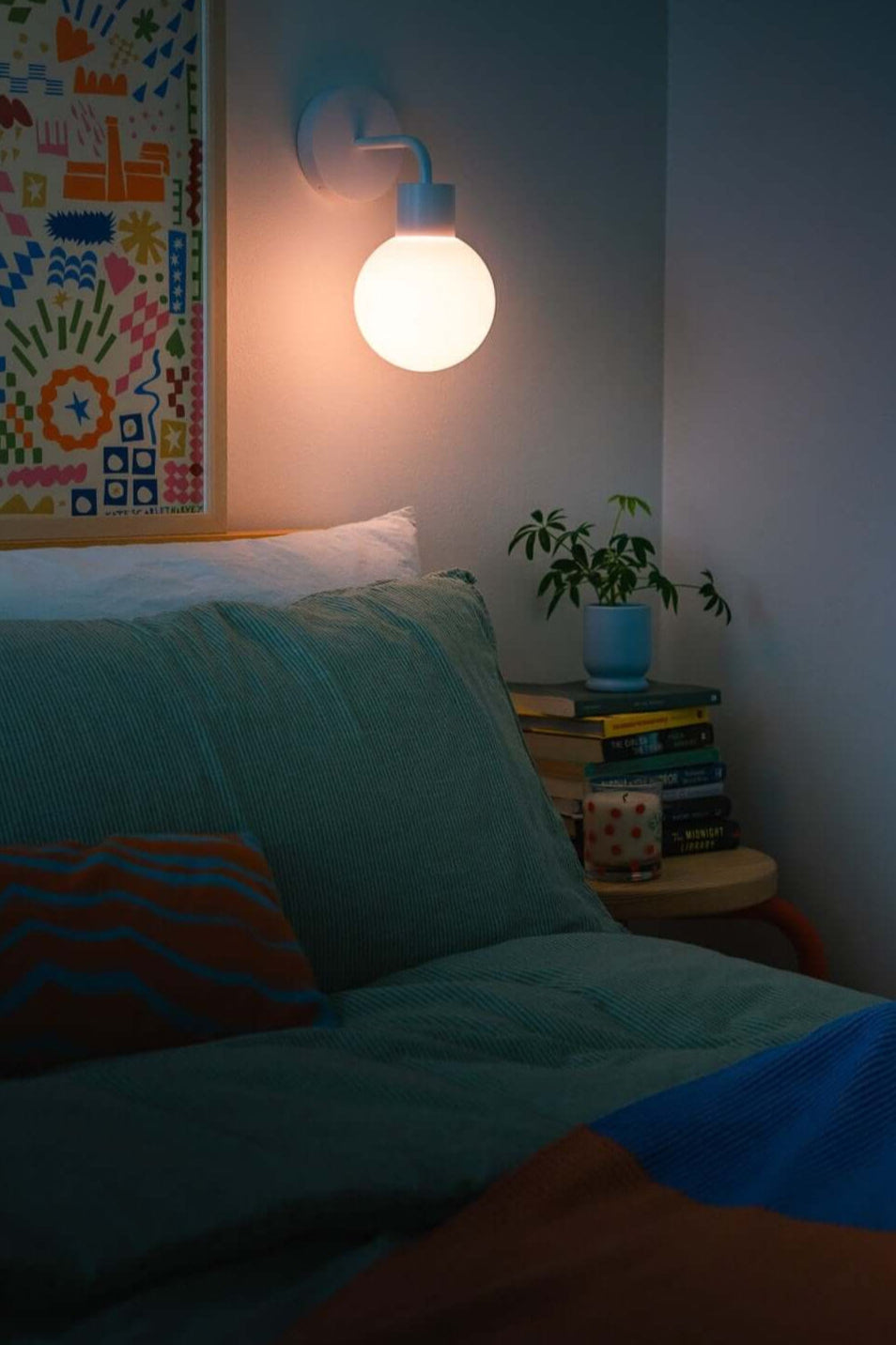 Cozy bedroom with a glowing wall lamp, colorful artwork, and a bedside plant and books, creating a serene night setting.
