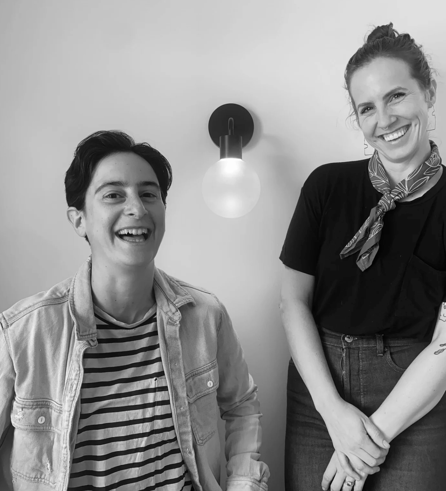 Two smiling individuals in a modern setting, one in a striped shirt and denim jacket, the other in a black top and scarf.