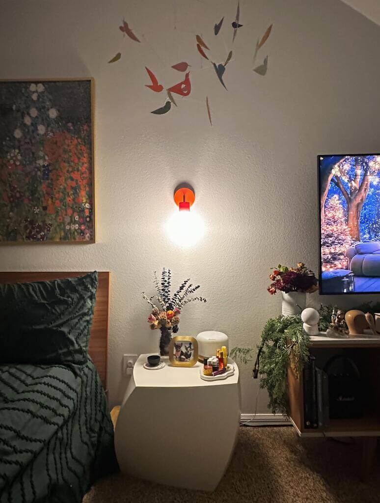 A vermillion red wall light sconce installed in a bedroom with a bedside table and some decorative artwork