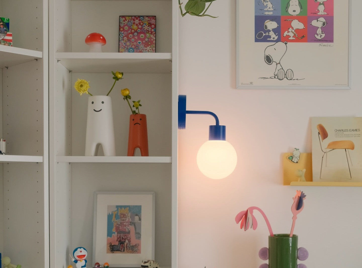 Cozy shelving unit displaying colorful art prints, cheerful vases with smiling faces holding yellow flowers, a Blue Poplight wall light sconce, and Snoopy artwork on the wall.