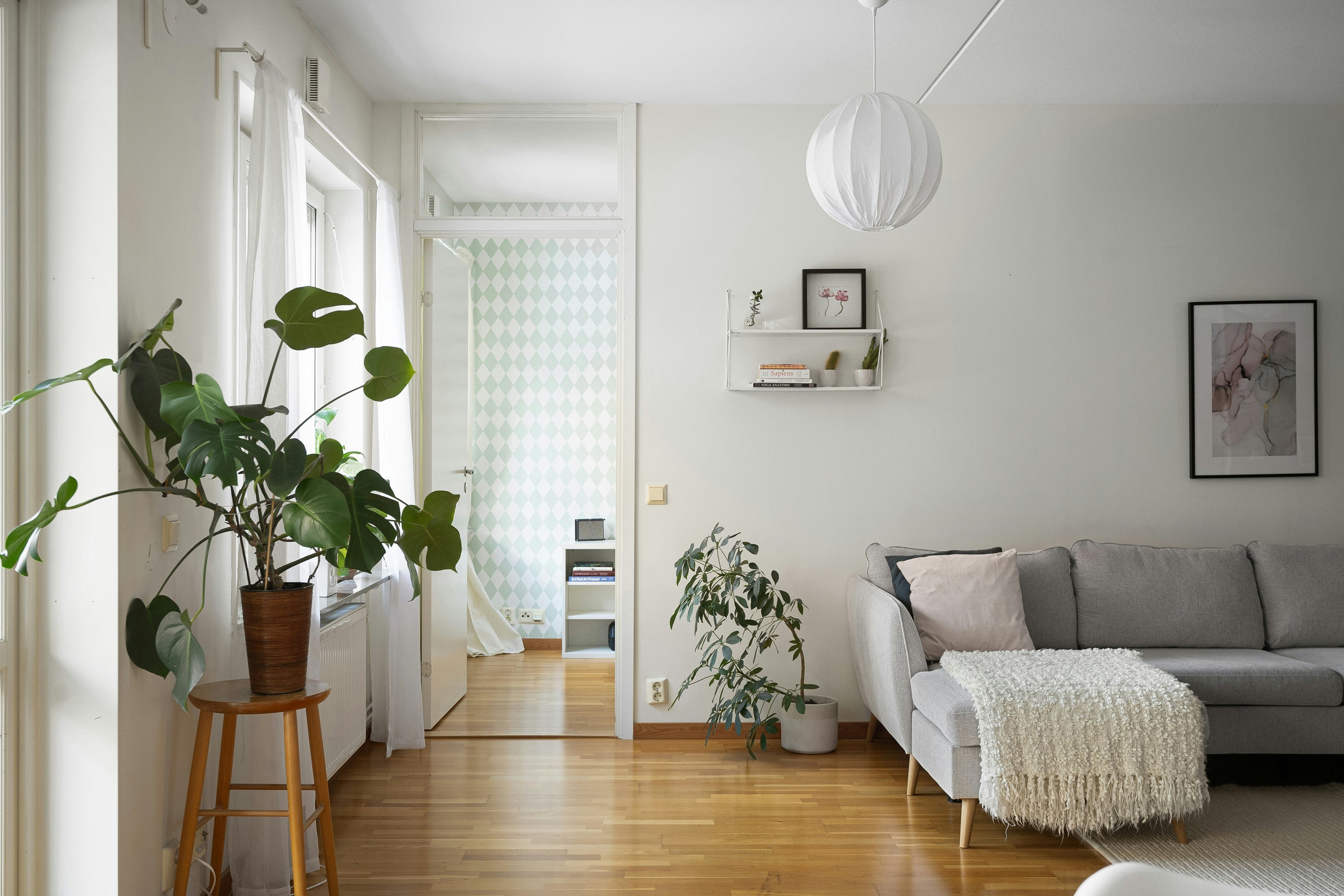 A minimalist Scandinavian-style living room with white walls and light wood flooring, featuring a gray sofa with white throw pillows and a fluffy white ottoman