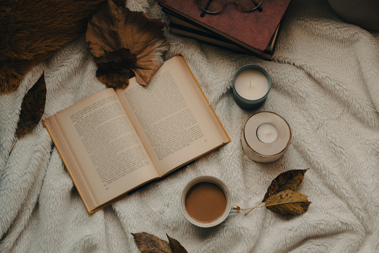 Flat lay of an open book on a cream knit blanket with a mug of coffee, two candles, dried autumn leaves, and reading glasses, creating a cozy fall reading atmosphere
