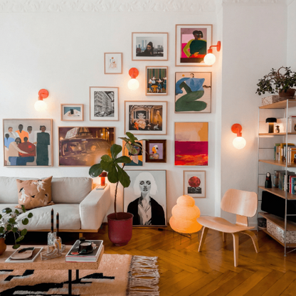 Living room with modern white sofa, gallery wall of framed art in natural wood frames, illuminated with Vermillion Red Poplight wall sconces, and wood herringbone flooring.
