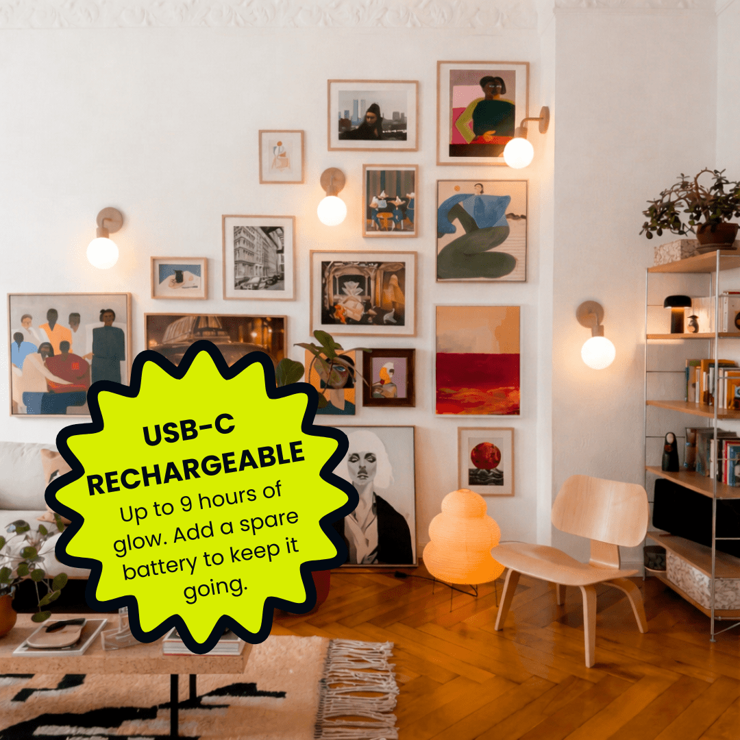 A cozy living room with an eclectic gallery wall of framed artwork and photographs in various sizes, warm wood flooring, and illuminated with Poplight Mushroom wall sconces with graphic overlay text "USB-C RECHARGEABLE up to 9 hours of glow."