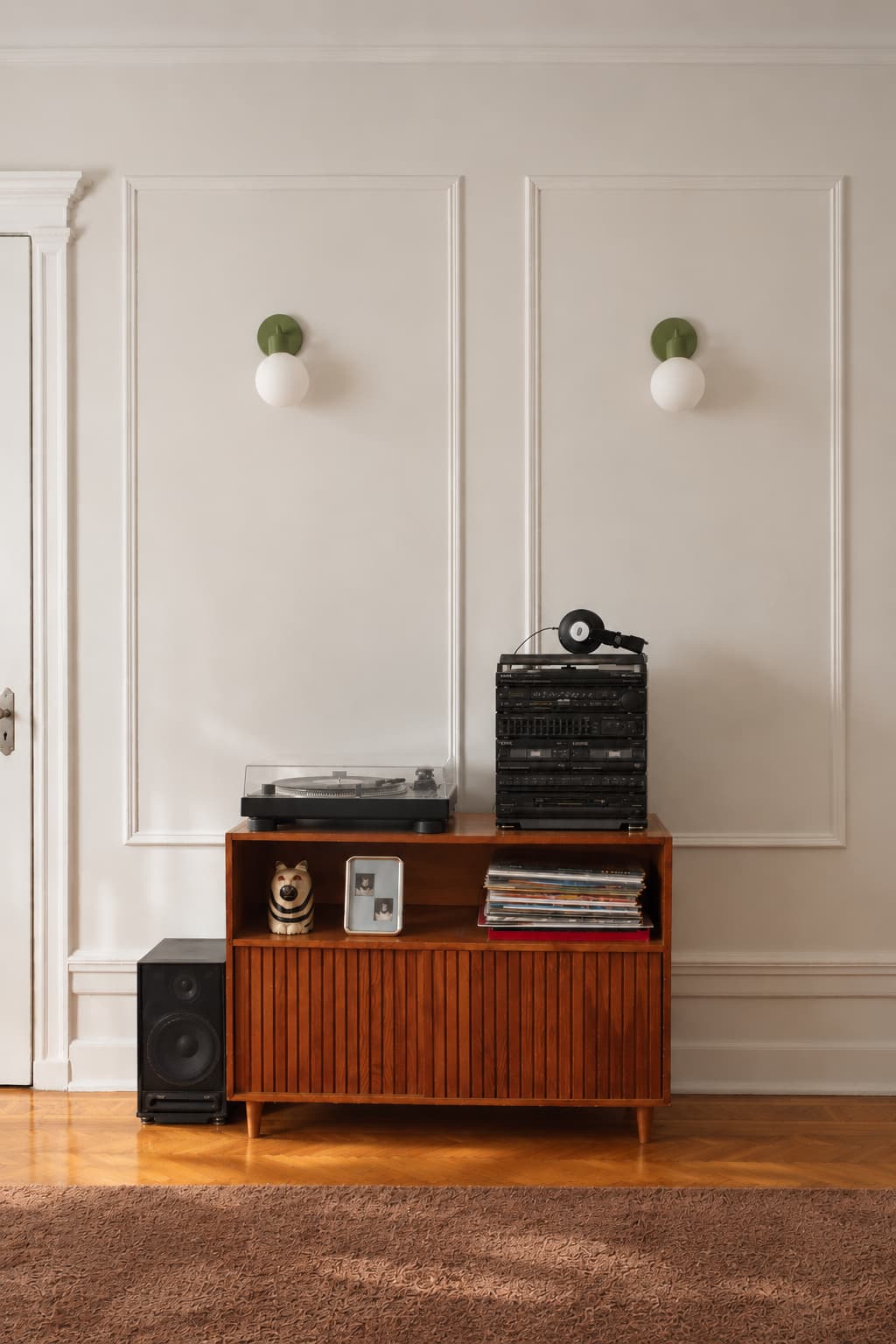 A mid-century modern wooden media console in a warm brown finish, styled with a turntable, a stack of CDs, a speaker, and records against a white paneled wall with two green and white wall sconces.