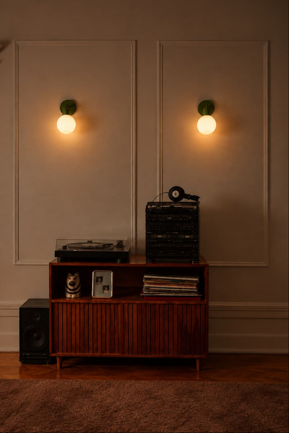 A warm, dimly lit interior scene featuring two green wall sconces mounted on a beige paneled wall above a wooden media console with a turntable and records. A brown rug is visible in the foreground.
