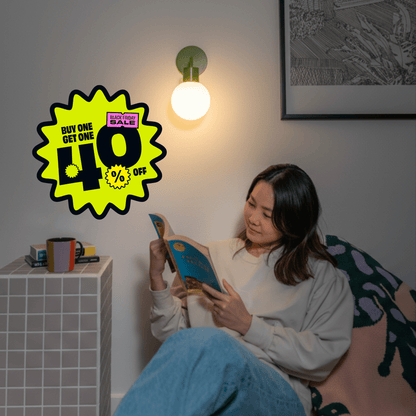 Woman reading a book in a cozy corner illuminated with a Sage Green Poplight wall light sconce and a promotional badge &