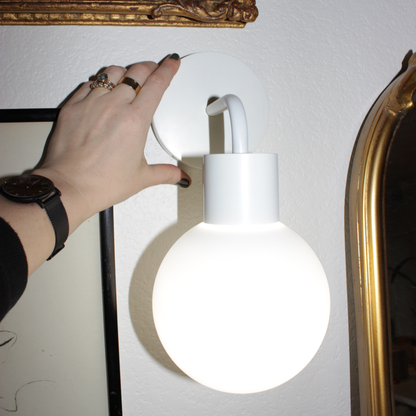 Hand adjusting modern white wall lamp with round bulb against textured wall and framed art.